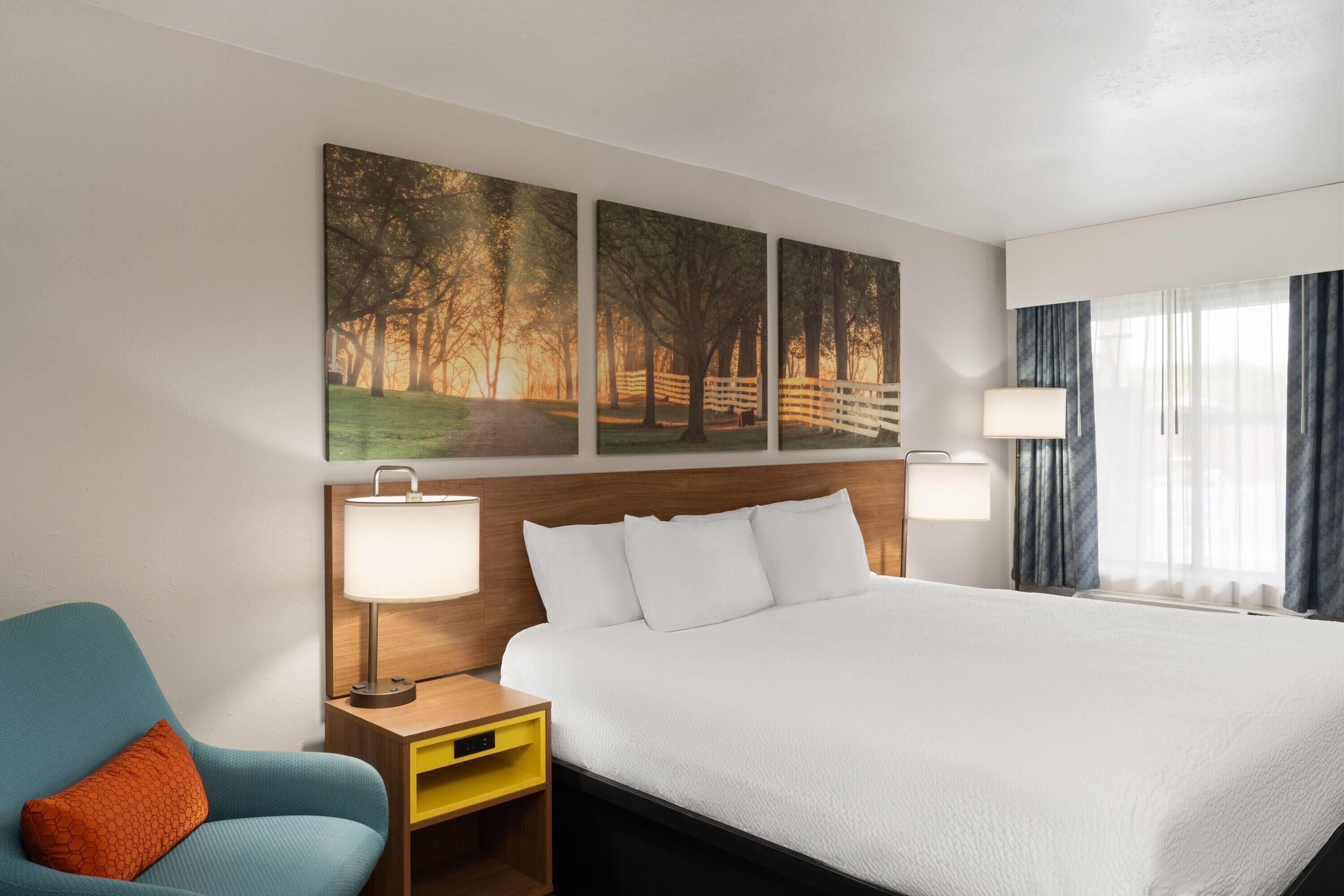 A hotel room with a large bed, two bedside lamps, a nightstand, a chair, and a painting of a forest scene.