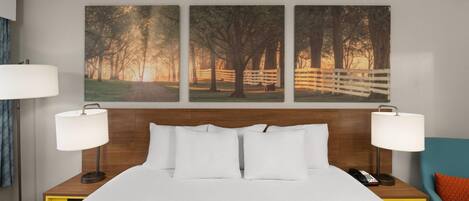 A hotel room with a bed, two bedside lamps, a nightstand, a chair, and three framed pictures on the wall.