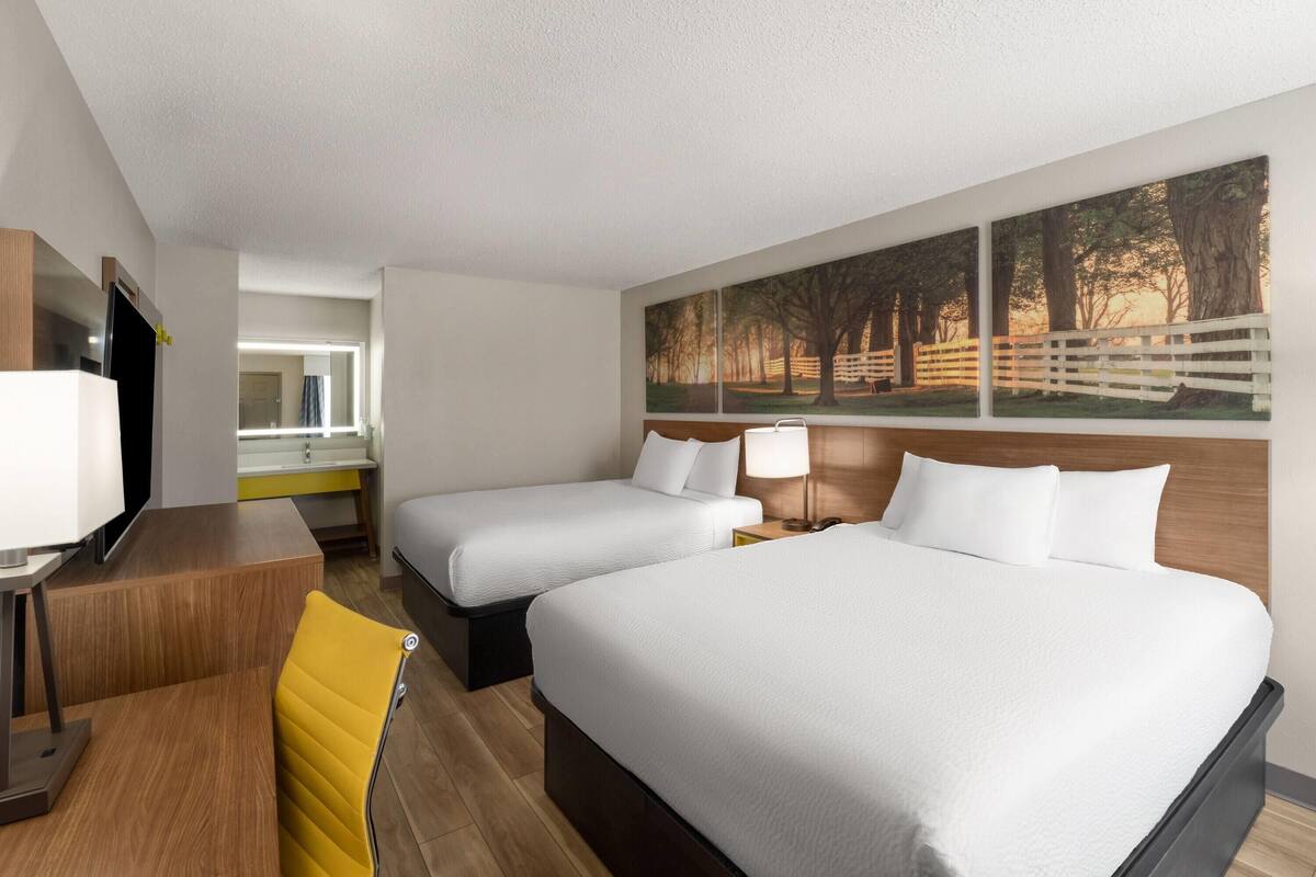 A hotel room with two beds, a desk, a chair, a TV, and a painting on the wall.