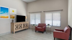 A living room with a TV, two windows with blinds, a red leather sofa, a wooden cabinet, and a small table with a plant. - Days Inn by Wyndham Clanton AL (Clanton)