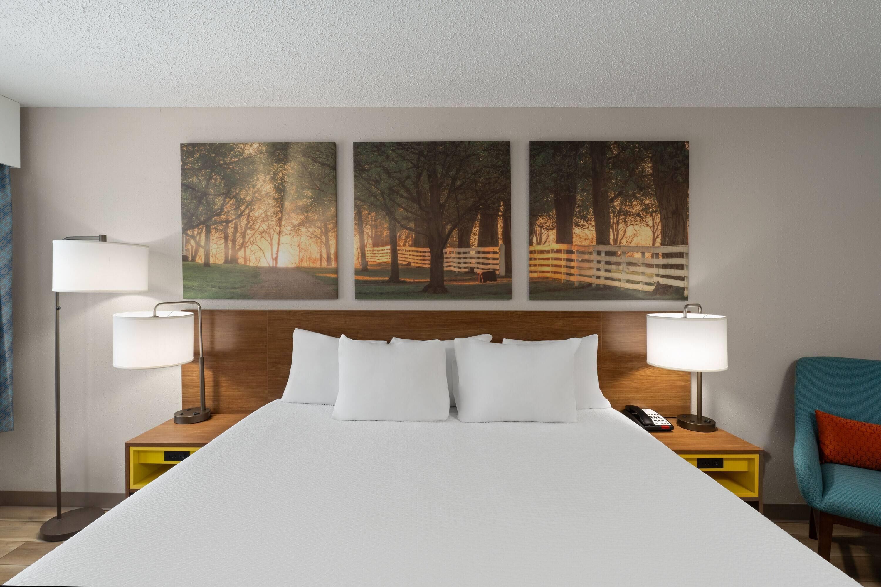 Ein Hotelzimmer mit einem großen Bett, zwei Nachttischlampen, einem hölzernen Kopfteil, einem Nachttisch mit Telefon, einem Sessel und drei gerahmten Bildern an der Wand.