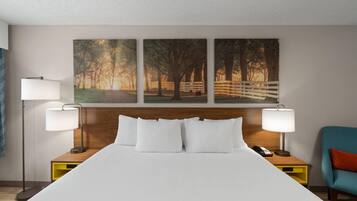 Ein Hotelzimmer mit einem großen Bett, zwei Nachttischlampen, einem hölzernen Kopfteil, einem Nachttisch mit Telefon, einem Sessel und drei gerahmten Bildern an der Wand.