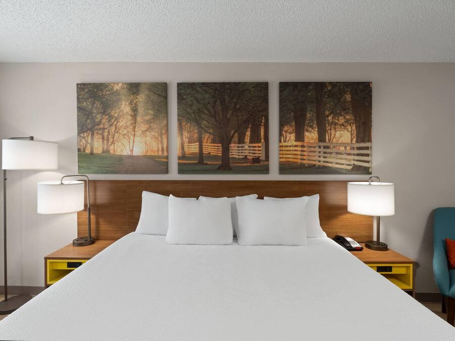 Ein Hotelzimmer mit einem großen Bett, zwei Nachttischlampen, einem hölzernen Kopfteil, einem Nachttisch mit Telefon, einem Sessel und drei gerahmten Bildern an der Wand.