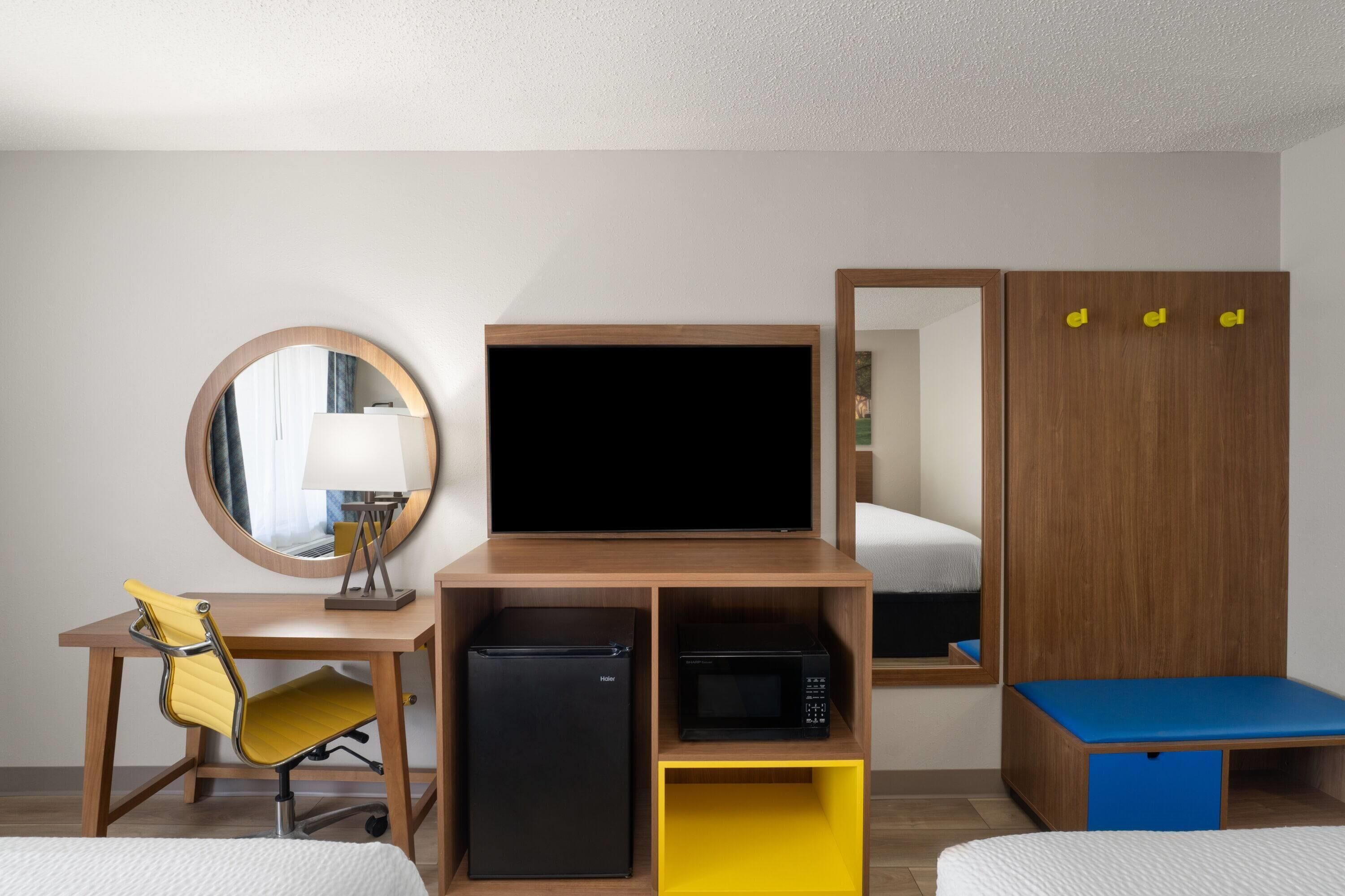 A hotel room with a desk, chair, television, refrigerator, microwave, and a bed with a blue mattress.