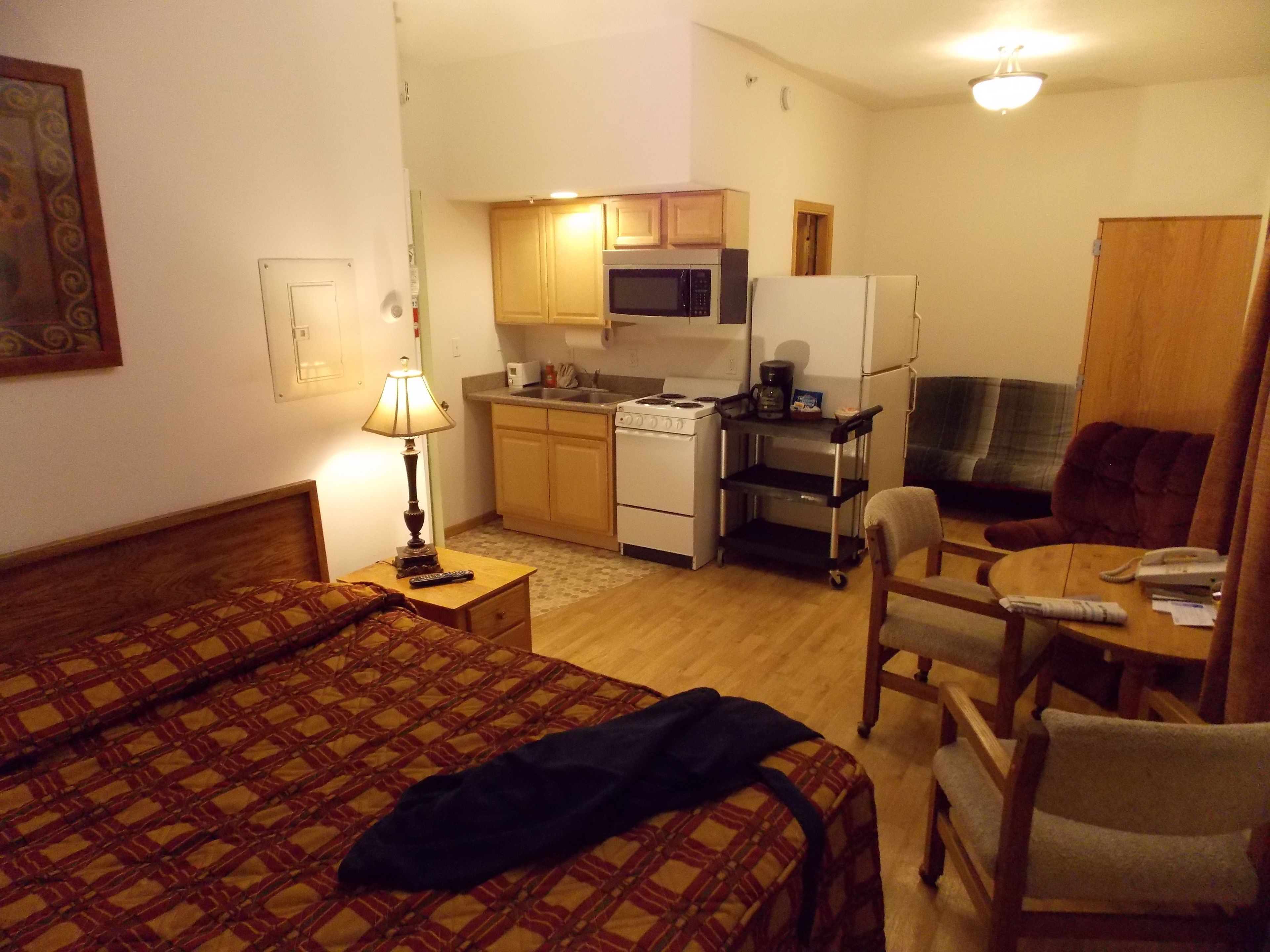 Ein kleines Hotelzimmer mit einem Bett, einem Schreibtisch mit Lampe, einer Küchenzeile mit Mikrowelle und Herd, einem Sessel und einem kleinen Tisch.