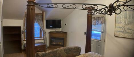 A bedroom with a four-poster bed, a television on a stand, a fireplace, and a window with curtains.
