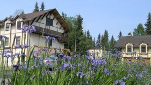A traditional chalet-style building with a sloped roof, surrounded by evergreen trees and a garden with purple flowers. - 7 Gables Inn & Suites (Fairbanks)