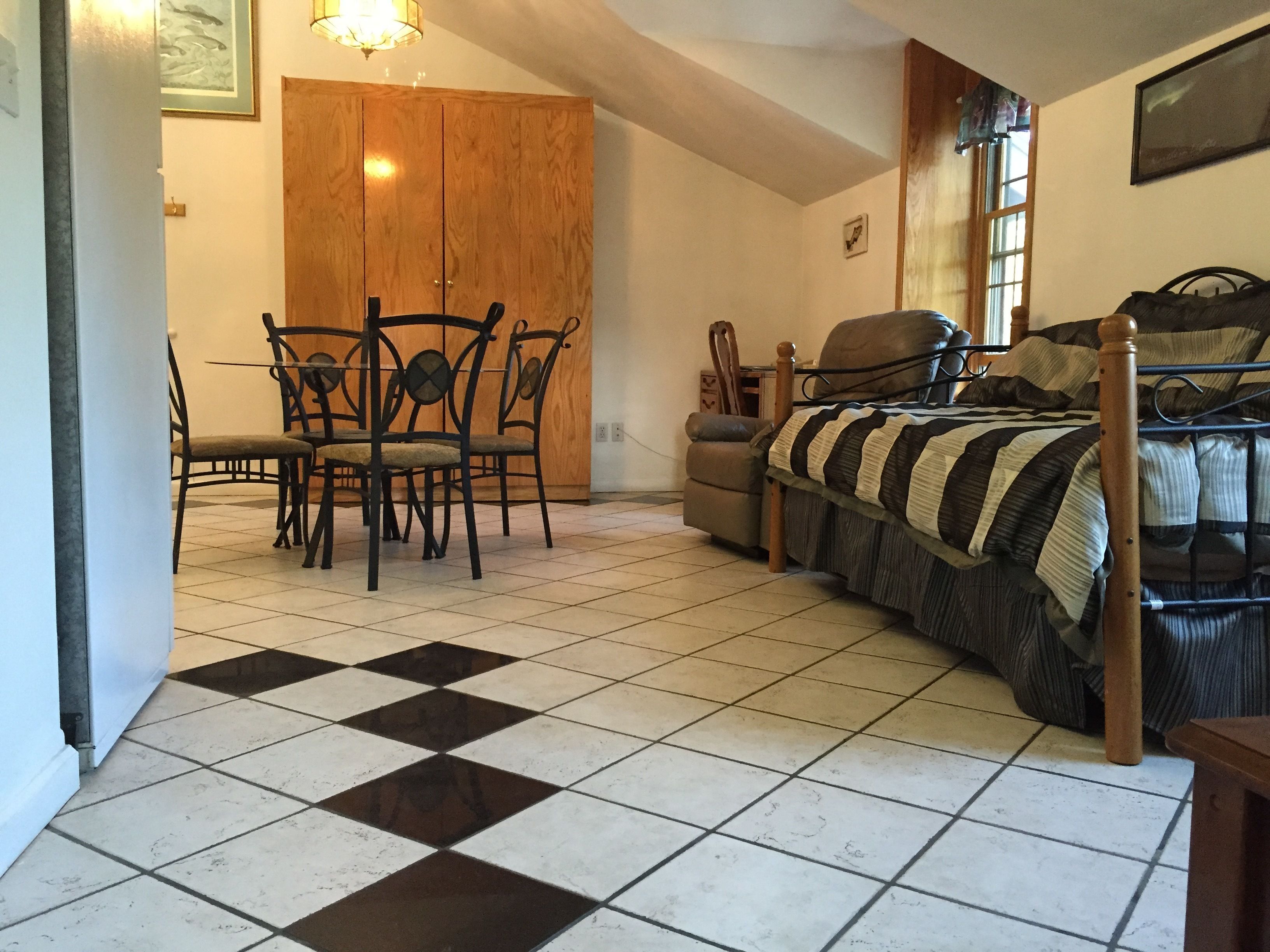 A room with a checkered floor, a bed with a striped blanket, a wooden wardrobe, a dining table with chairs, and a small table with a chair.