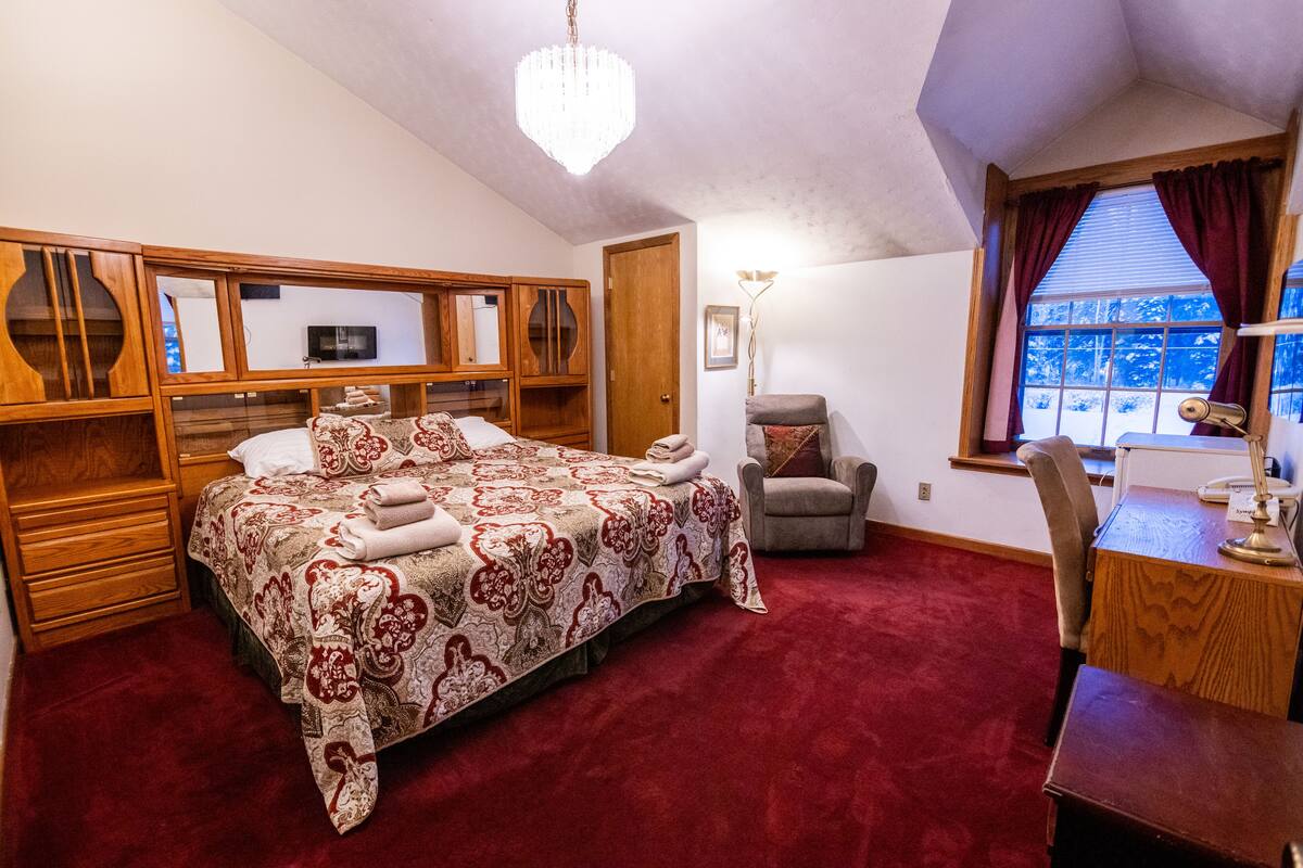 Une chambre à coucher avec un grand lit, une tête de lit en bois, un bureau avec une chaise, une fenêtre avec des rideaux et un lustre.
