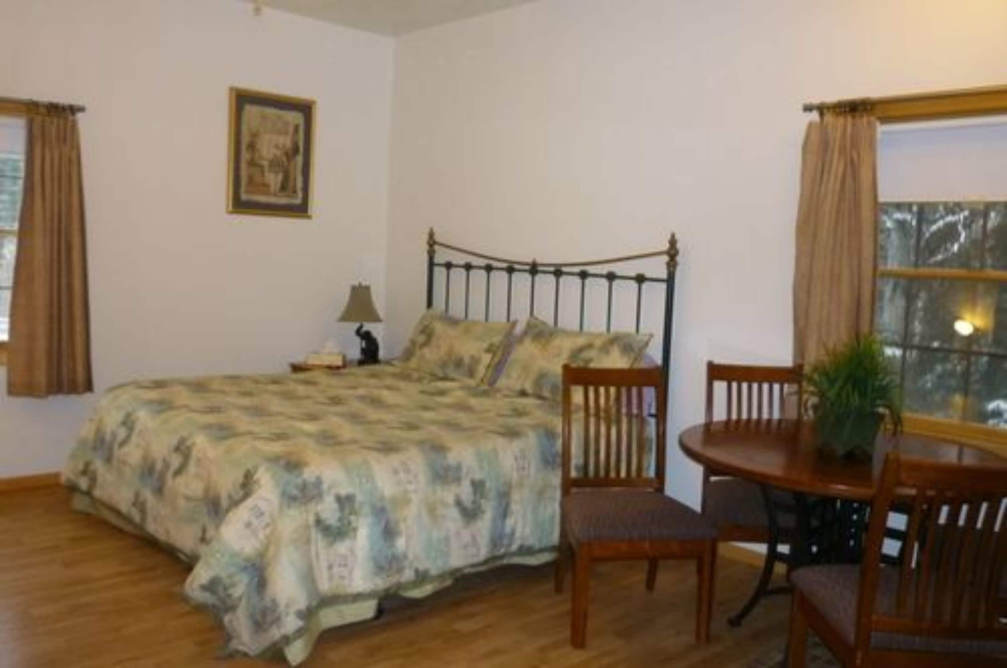 Un dormitorio con cama, mesita de noche, lámpara, silla, mesa de comedor y ventana con cortinas.