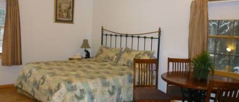 Un dormitorio con cama, mesita de noche, lámpara, silla, mesa de comedor y ventana con cortinas.