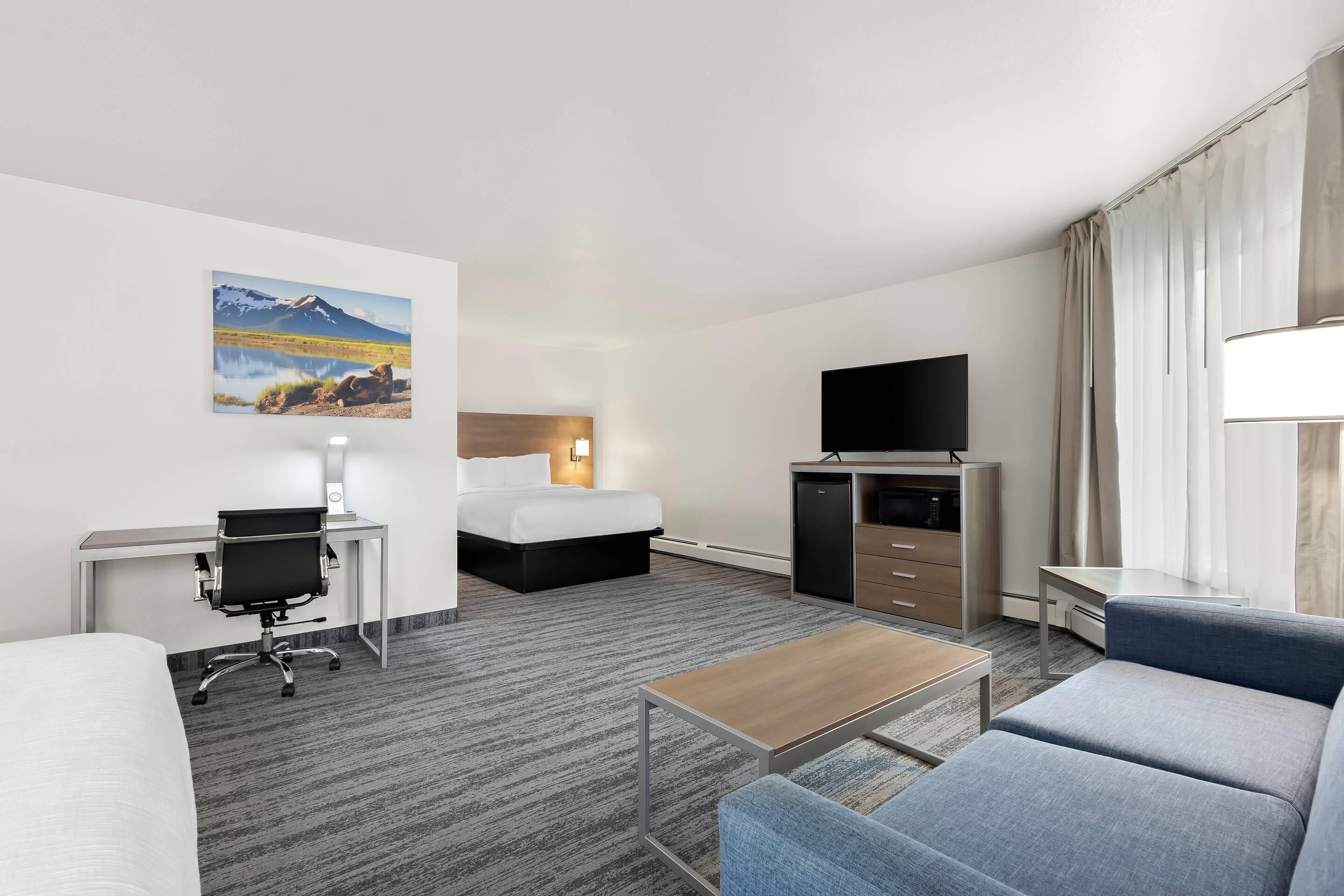 A hotel room with a bed, a desk with a chair, a TV, a sofa, a coffee table, and a painting of a mountain landscape.