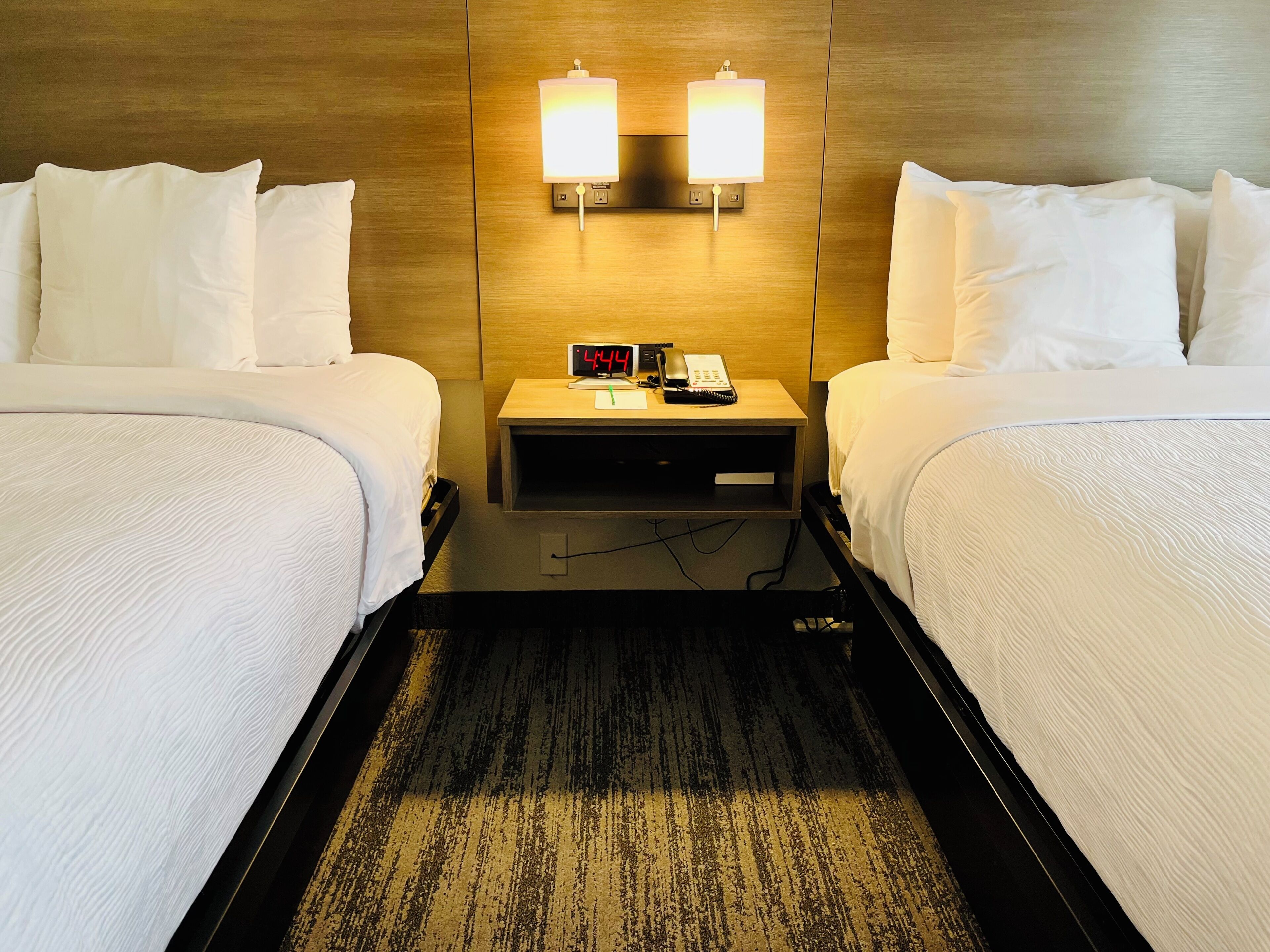 Habitación de hotel con dos camas, una mesita de noche con reloj y teléfono, y lámparas fijadas a la pared.