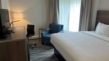 Hotellihuone, jossa on suuri sänky, työpöytä, jossa on tietokone, tuoli ja ikkuna, jossa on verhot.