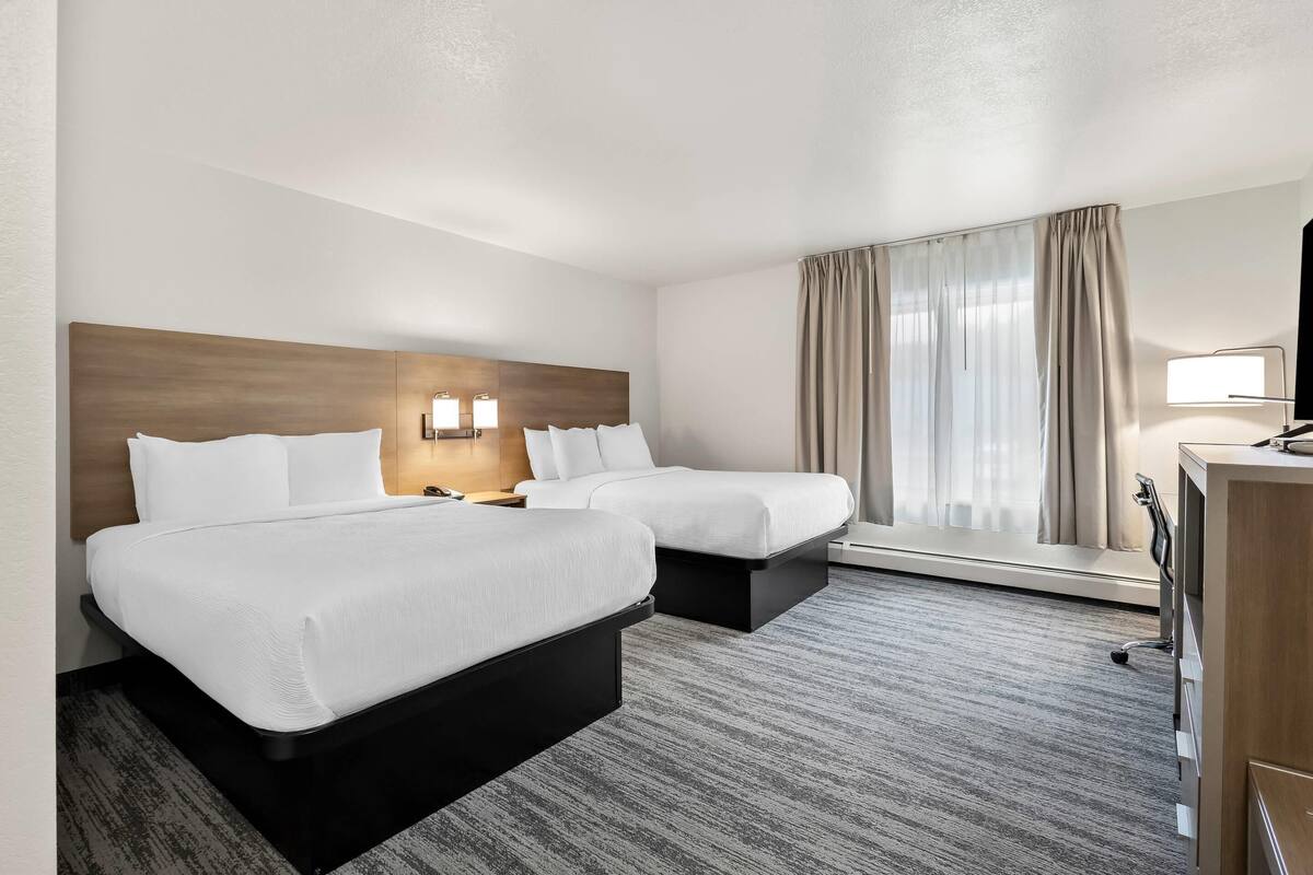 A hotel room with two beds, a desk, a lamp, and a window with curtains.