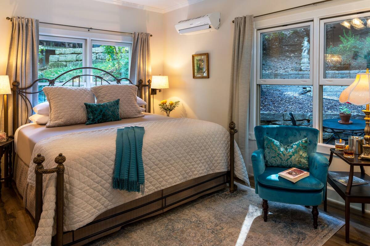 A bedroom with a bed, a chair, a table, and a window with a view of greenery.