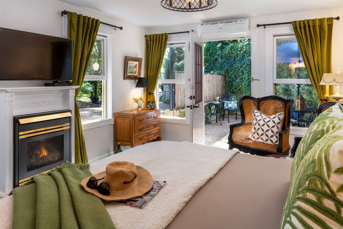 A bedroom with a fireplace, a television, a bed with a green blanket, a hat, and a chair with a patterned pillow.