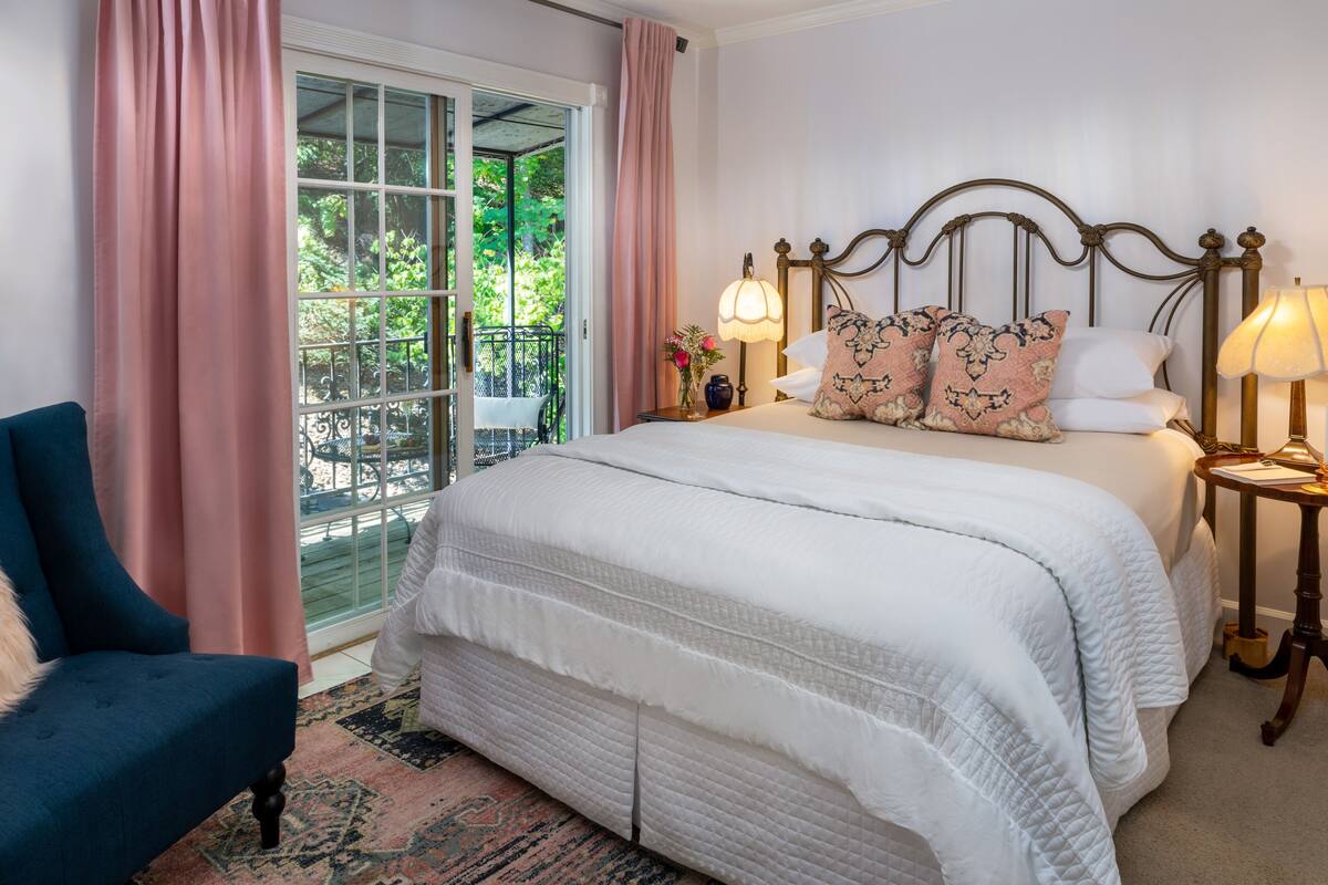 A bedroom with a large bed, two bedside lamps, a chair, and a view of greenery through a glass door.