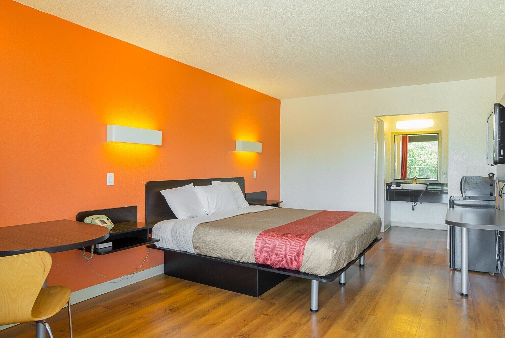 A hotel room with a bed, a desk, a chair, a small table, and a bathroom with a sink and mirror.