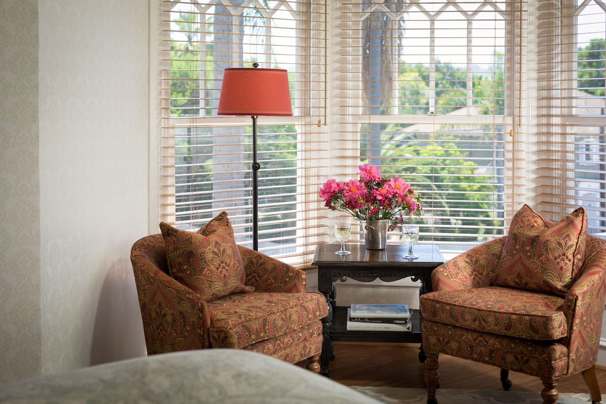 A room with a bed, two patterned armchairs, a small table with a vase of flowers, a lamp, and a window with blinds.