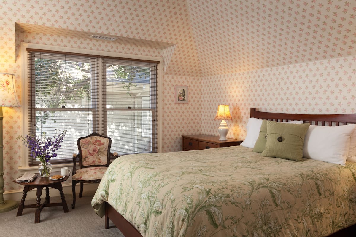 A bedroom with a floral-patterned bedspread, a wooden nightstand with a lamp, a chair, and a window with blinds.
