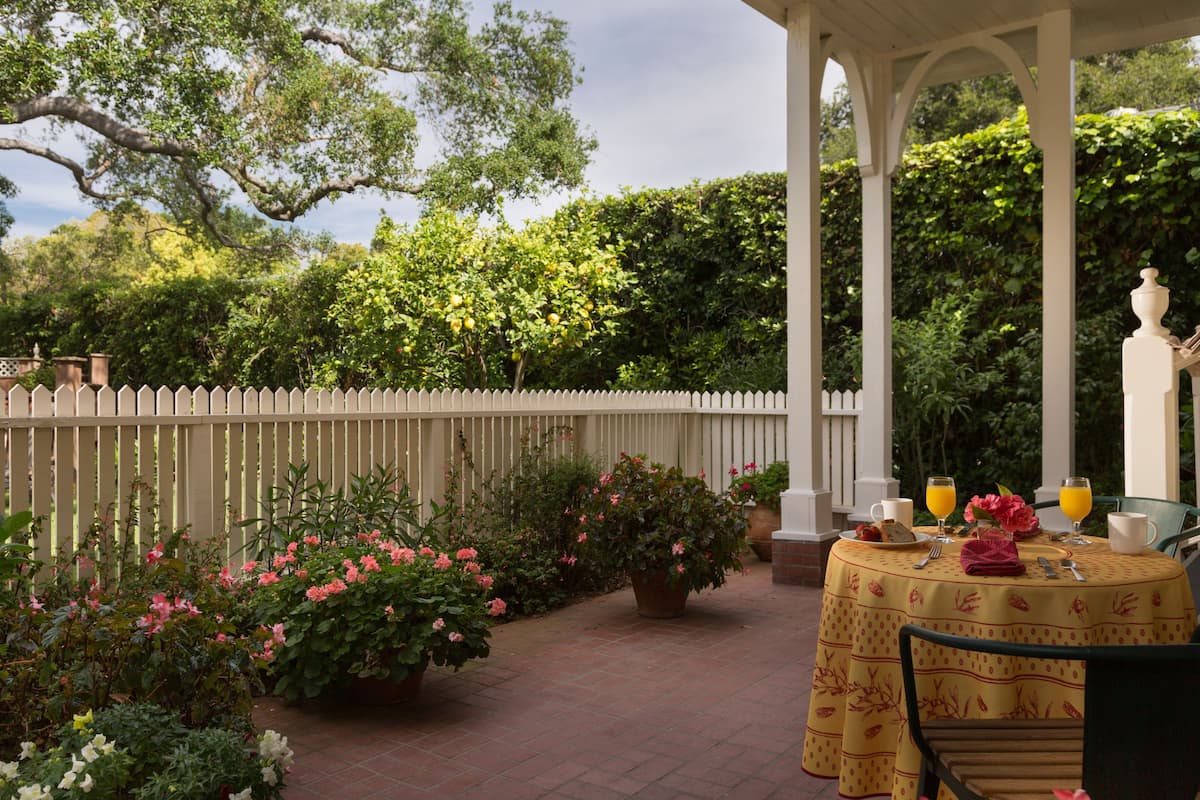 A covered patio with a table set for breakfast, surrounded by potted plants and a white picket fence.