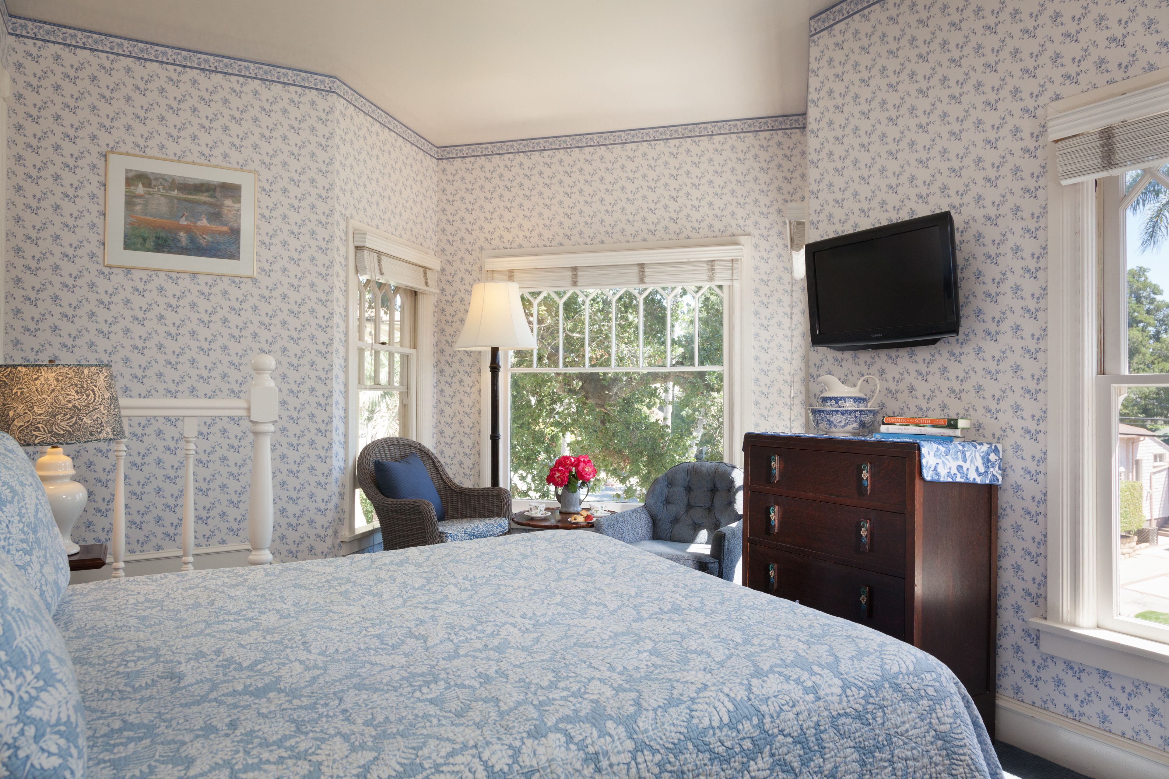 A bedroom with floral wallpaper, a bed with a blue and white patterned bedspread, a dresser, a television, and a window with a view of trees.