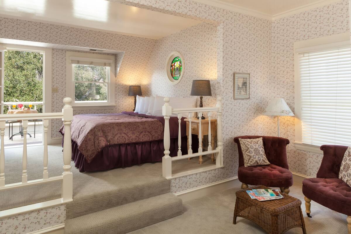 A bedroom with a bed, two chairs, a small table, a window, and a staircase.