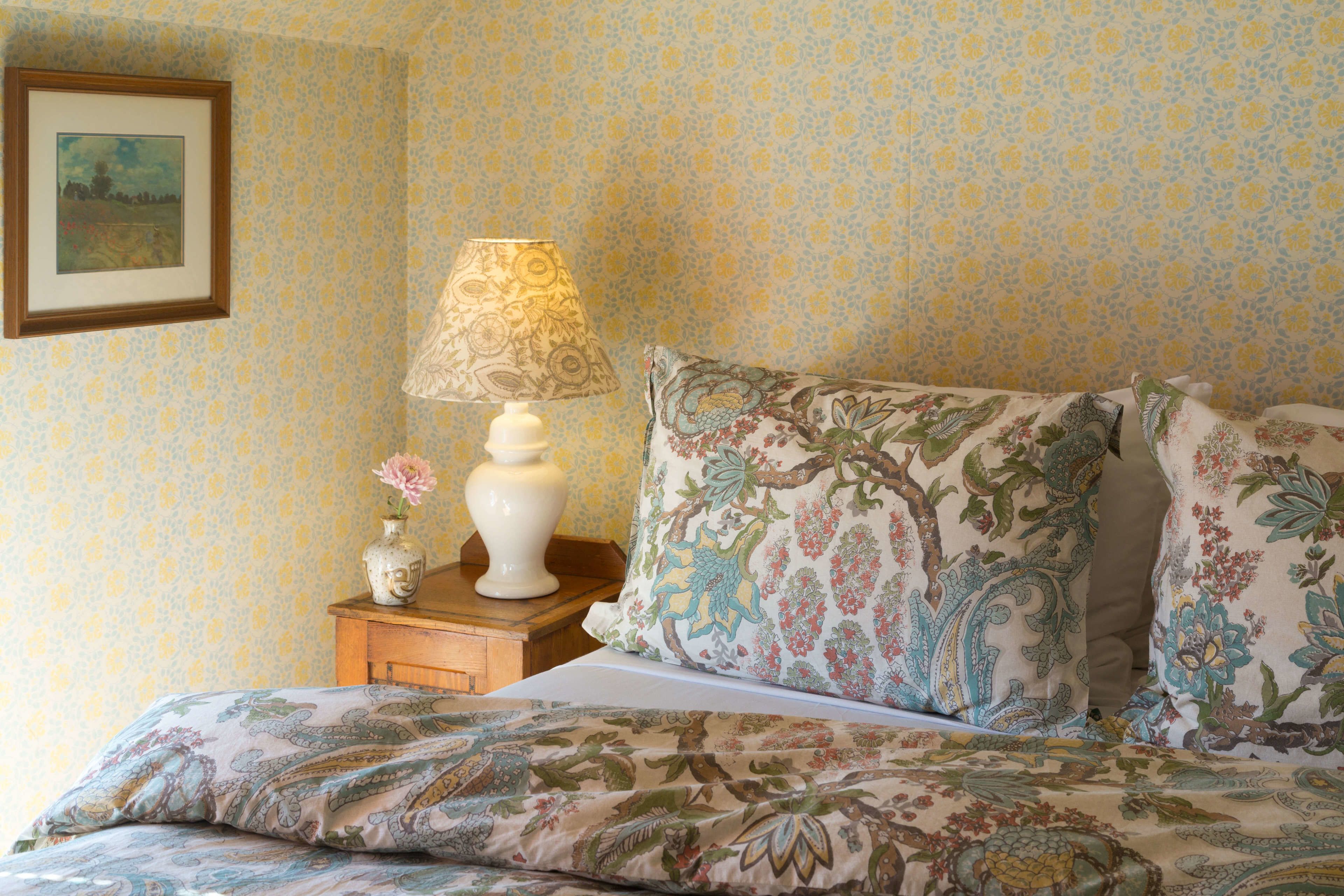 A bedroom with floral patterned bedding, a bedside table with a lamp, and a framed picture on the wall.