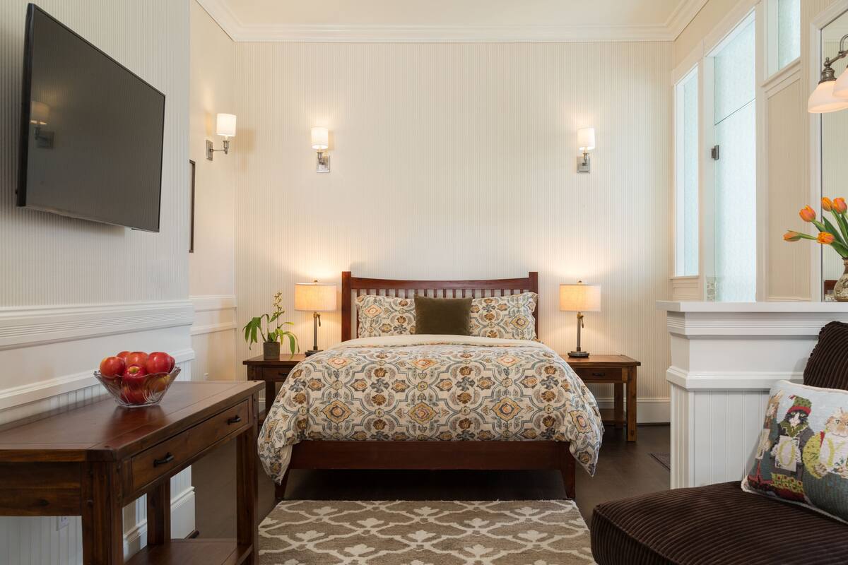A hotel room with a bed, bedside lamps, a TV mounted on the wall, a wooden nightstand, a small table with a bowl of fruit, and a window with a view of the outside.