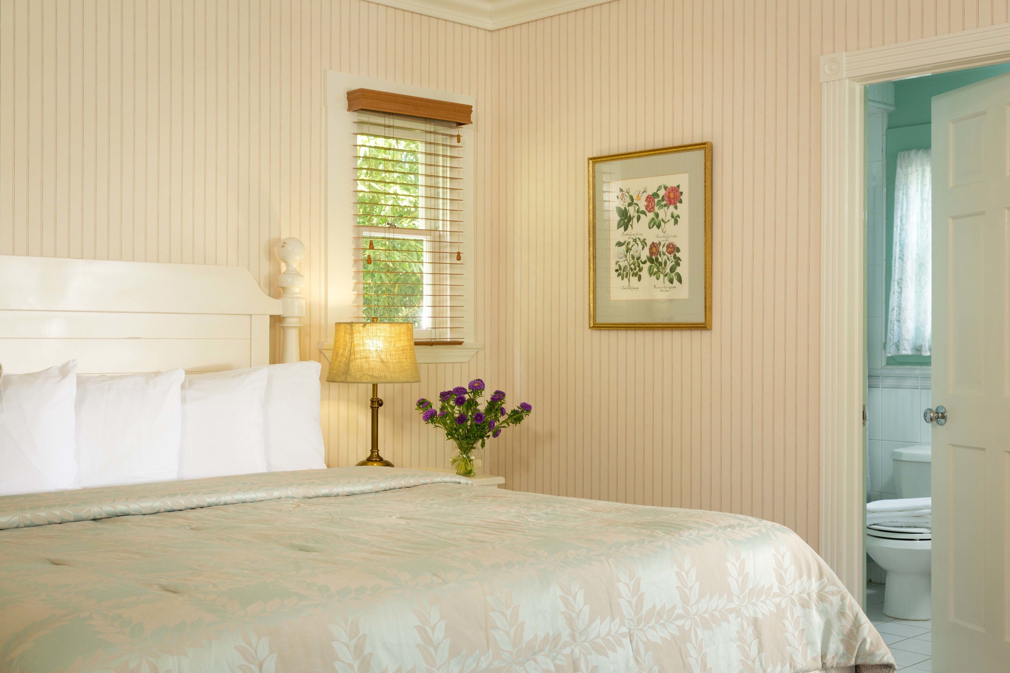 A neatly arranged bedroom with a bed, a bedside lamp, a framed botanical print, and a window with blinds.