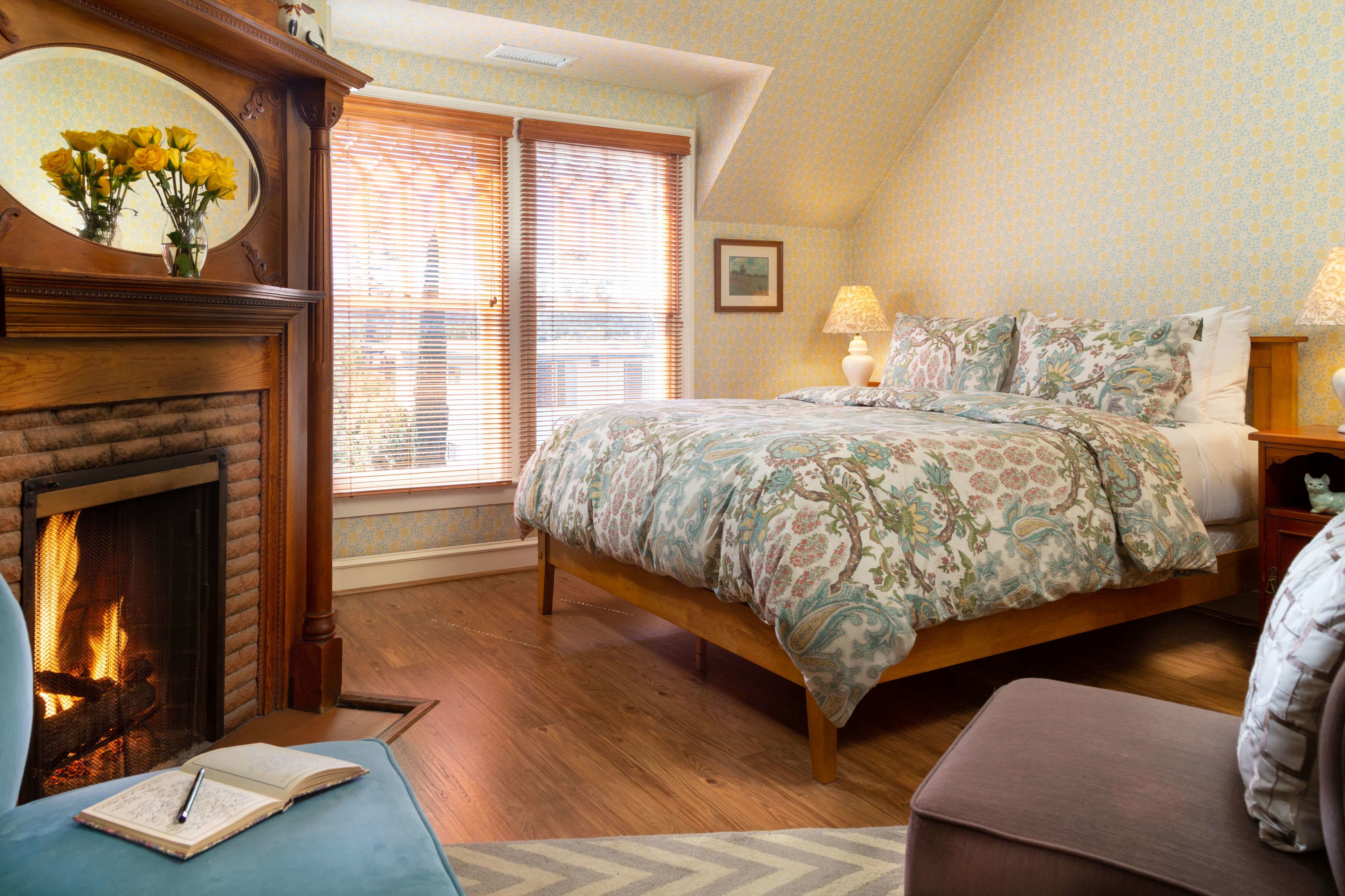 A bedroom with a fireplace, a bed with floral bedding, a nightstand with a lamp, and a window with blinds.