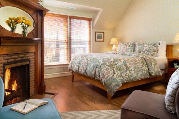 A bedroom with a fireplace, a bed with floral bedding, a nightstand with a lamp, and a window with blinds.