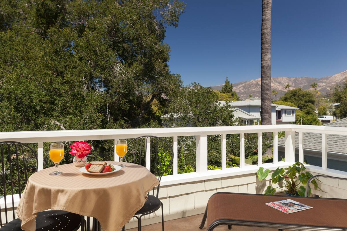 A balcony with a table set for breakfast, including a plate of food, a glass of orange juice, and a flower. There is a chair and a magazine on the table.