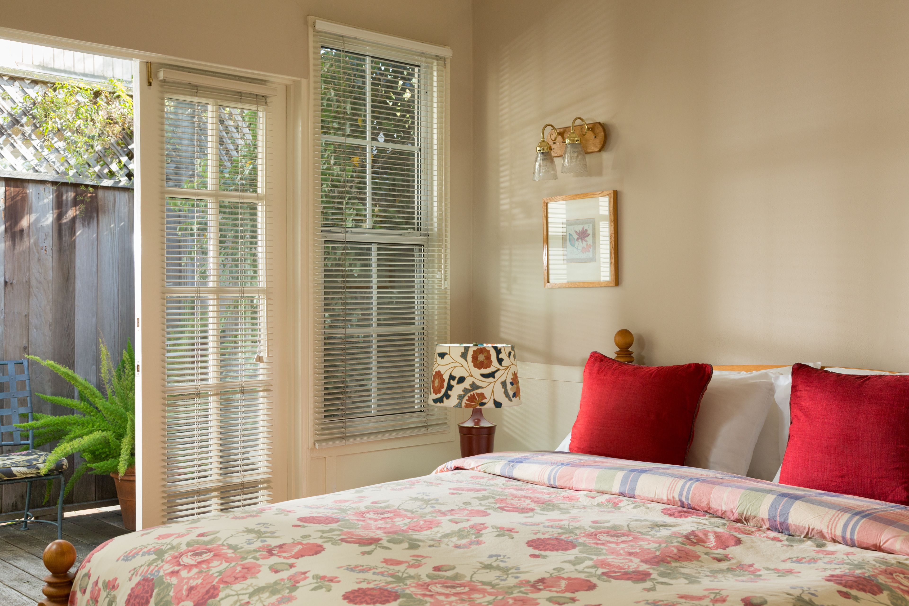 A bedroom with a floral bedspread, a wooden headboard, a nightstand with a lamp, and a window with blinds.