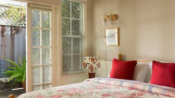 A bedroom with a floral bedspread, a wooden headboard, a nightstand with a lamp, and a window with blinds.