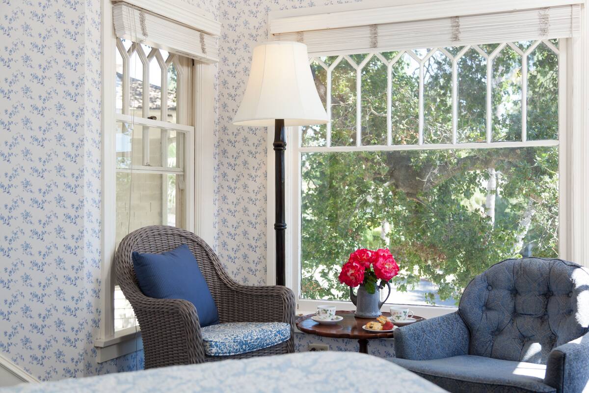 A room with a wicker chair, a blue upholstered chair, a table with a vase of flowers, and a window with a view of trees.