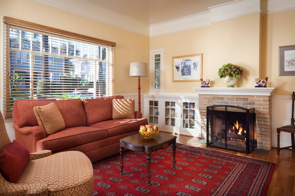 A living room with a red sofa, patterned ottoman, and a fireplace with a fire.
