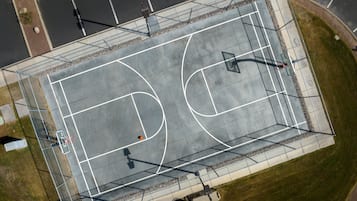 An aerial view of a basketball court with a fence, basketball hoops, and a parking lot in the background.