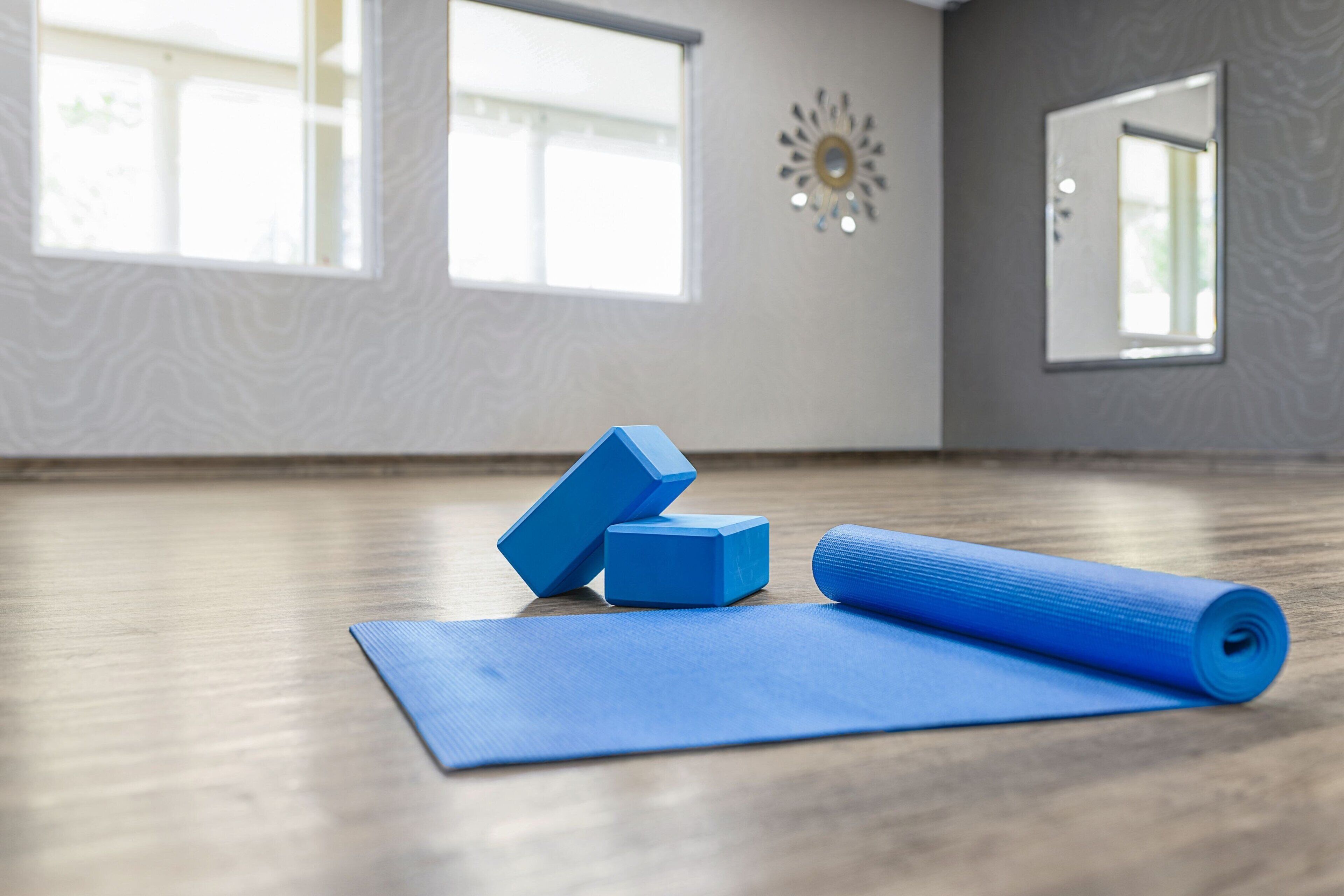 Eine Yogamatte, zwei Schaumstoffrollen und ein Yoga-Block auf einem Holzboden in einem Raum mit großen Fenstern und einem Spiegel.