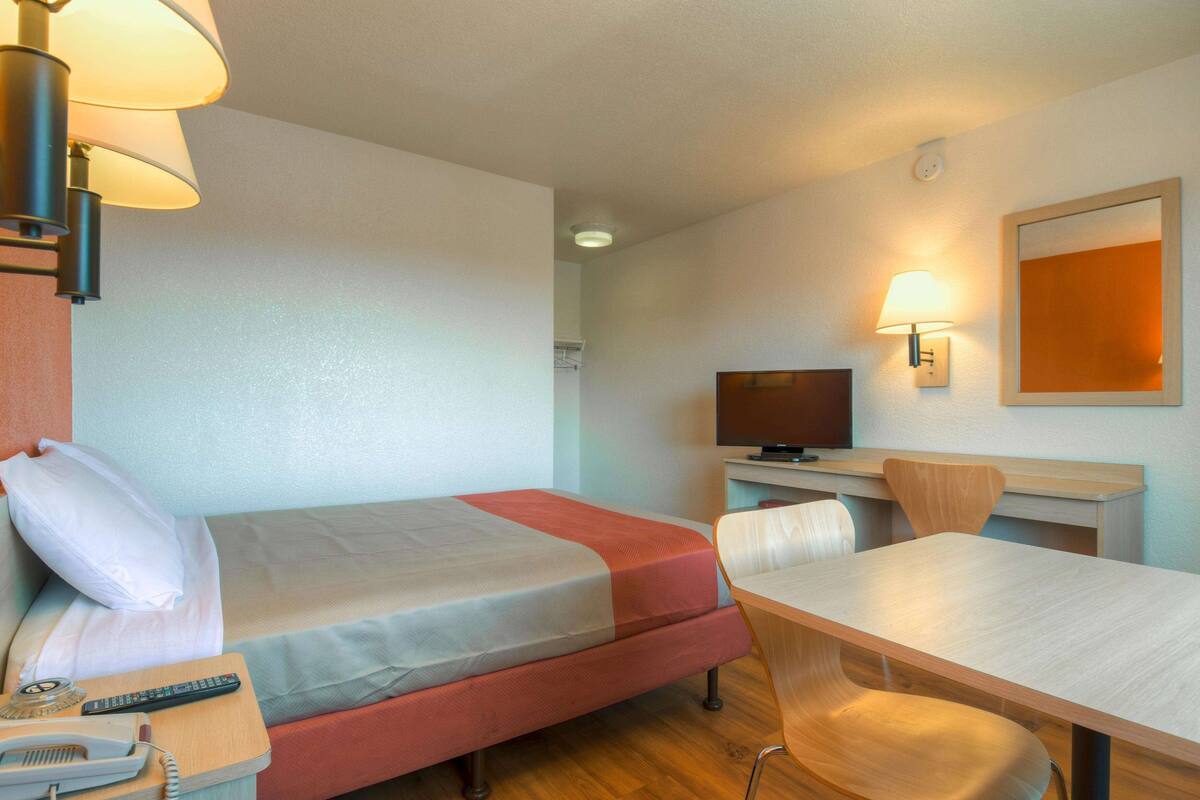 A hotel room with a bed, bedside table, lamp, TV, desk, chair, and a telephone.