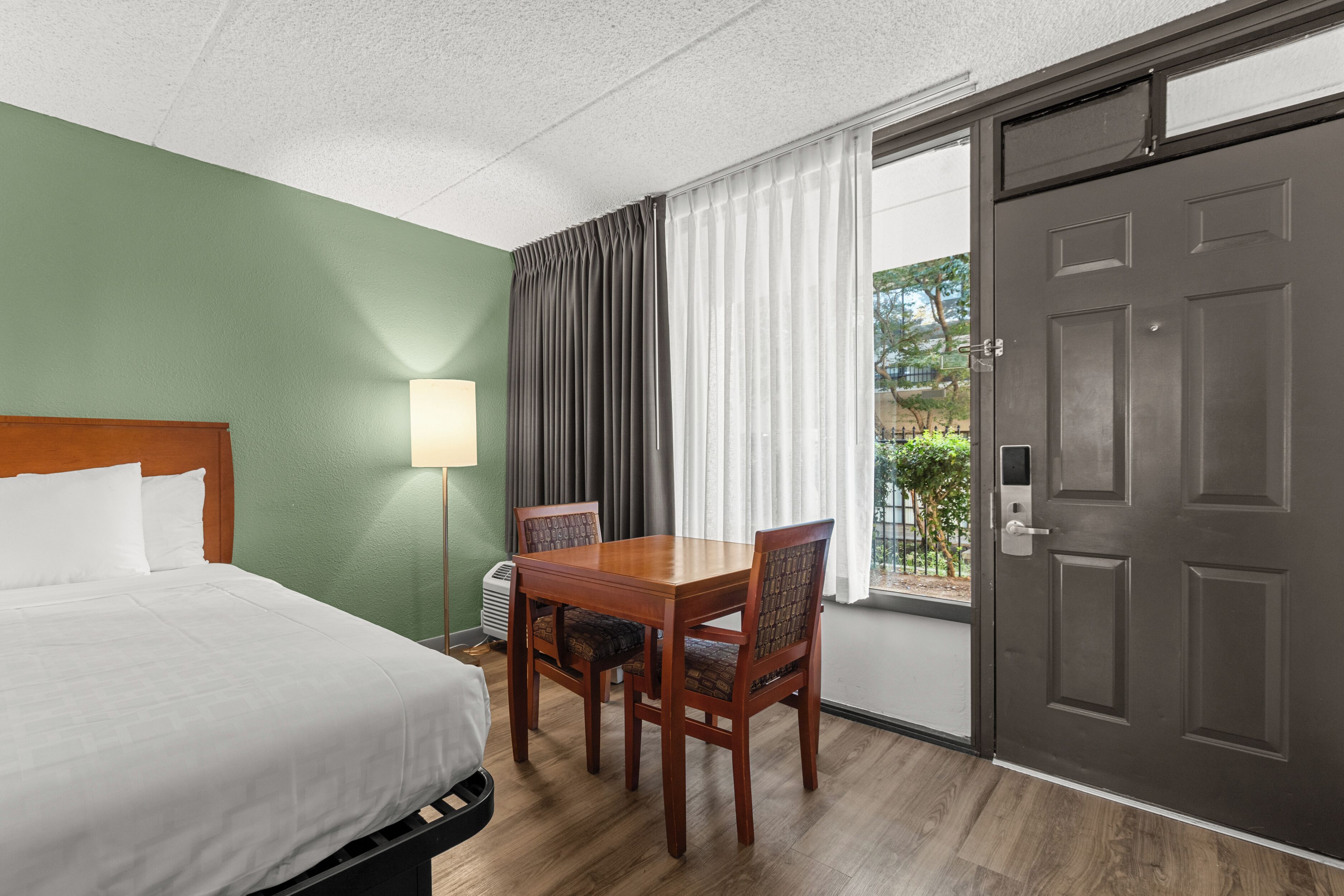 A hotel room with a bed, a table with chairs, a lamp, and a door.