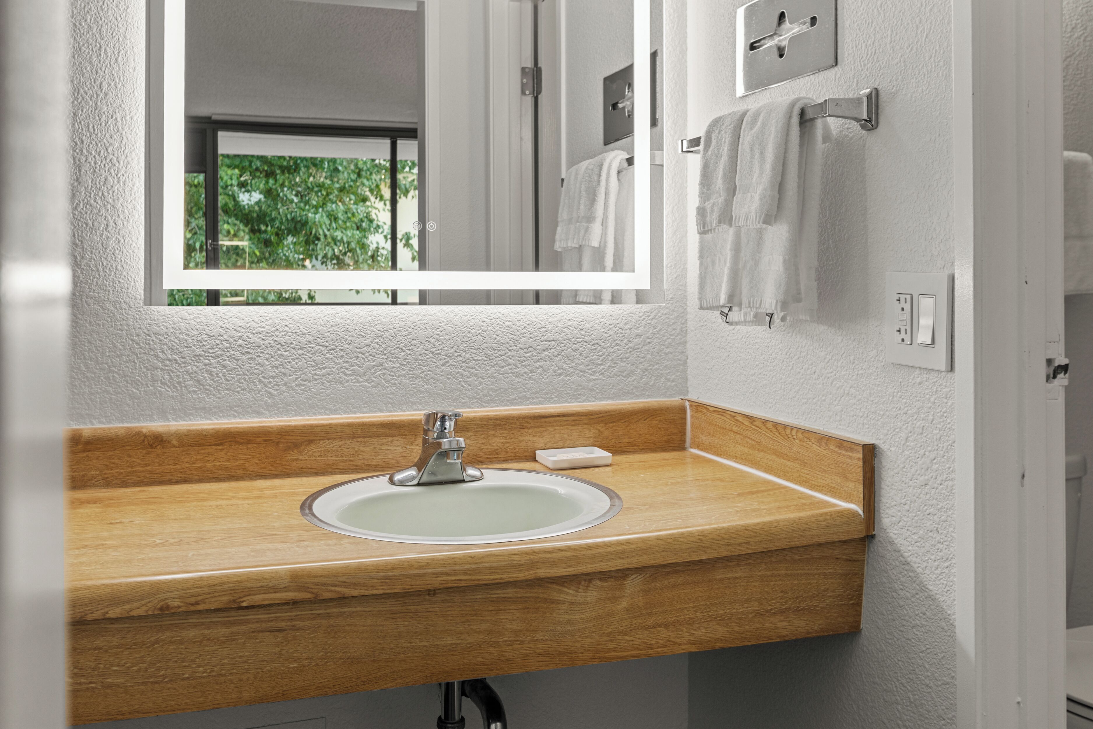 A bathroom with a wooden vanity, a white sink, a mirror, and a window with a view of greenery.