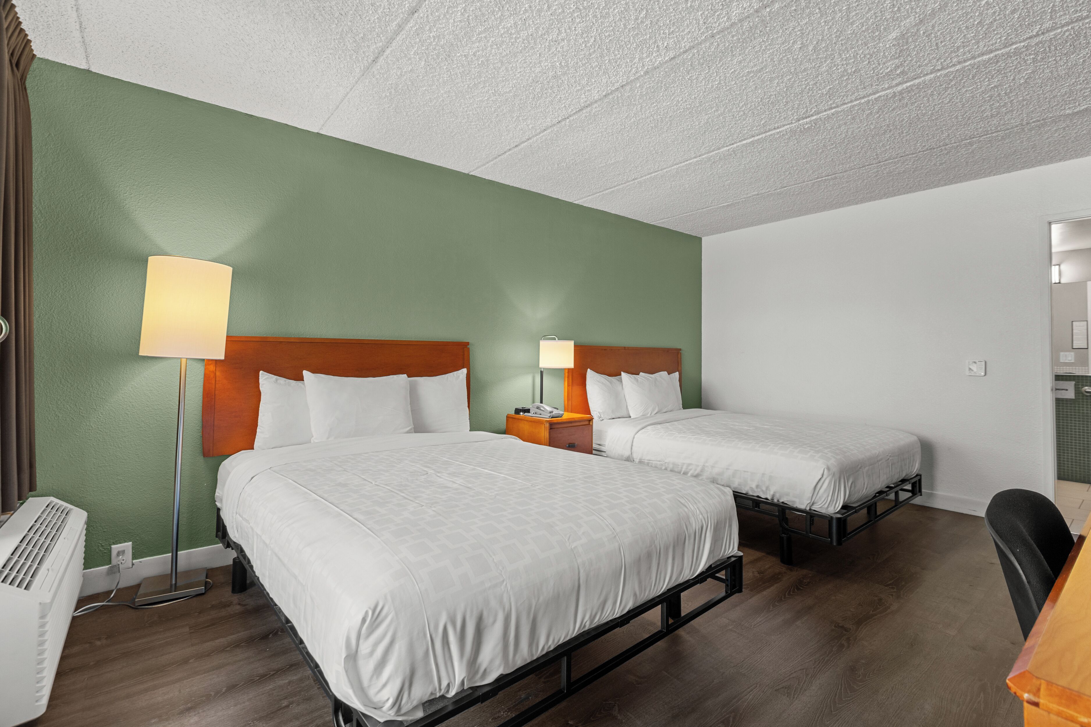 A hotel room with two beds, a desk, a chair, a lamp, and a heater.
