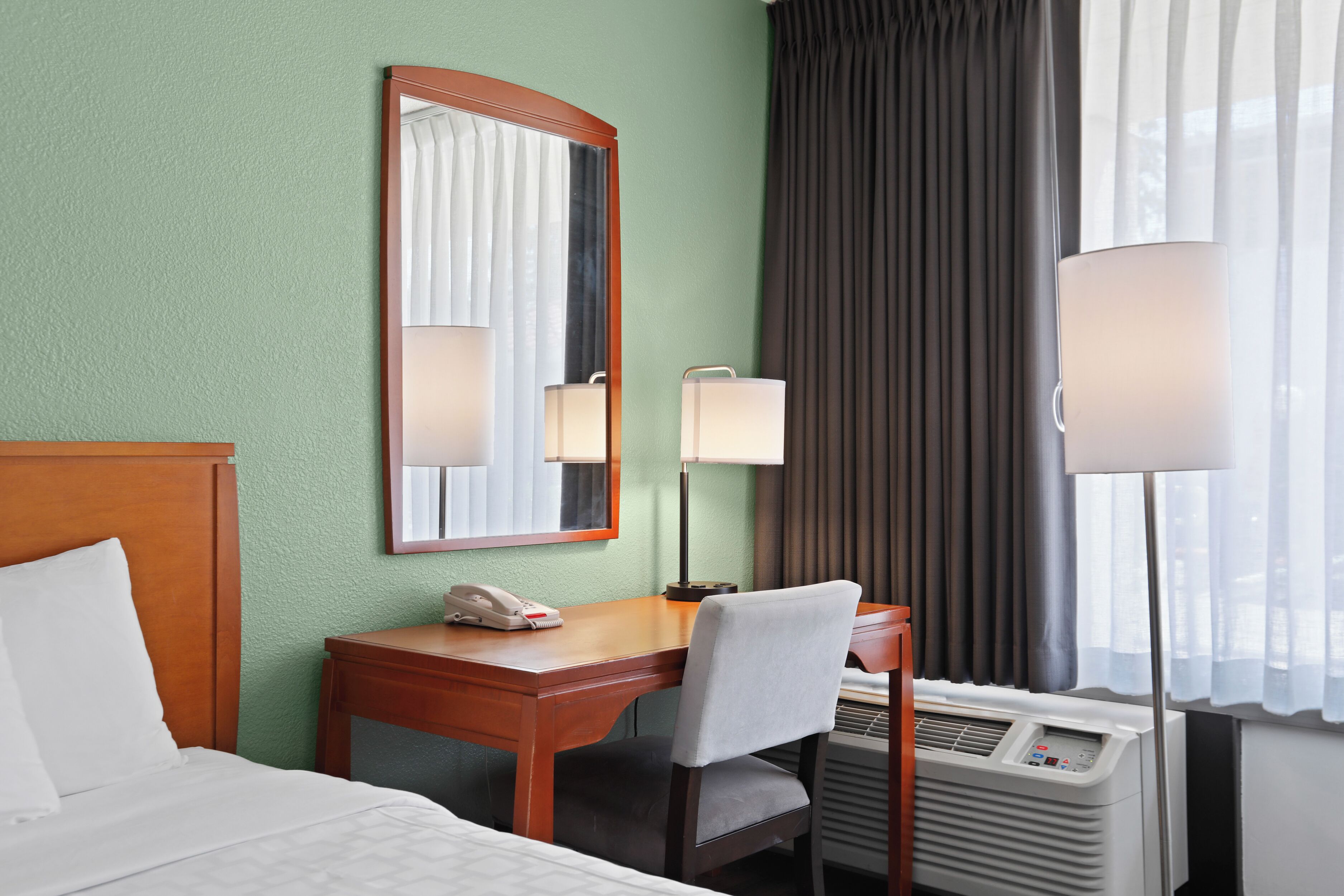 A hotel room with a bed, a desk, a chair, a telephone, a mirror, a lamp, and an air conditioning unit.
