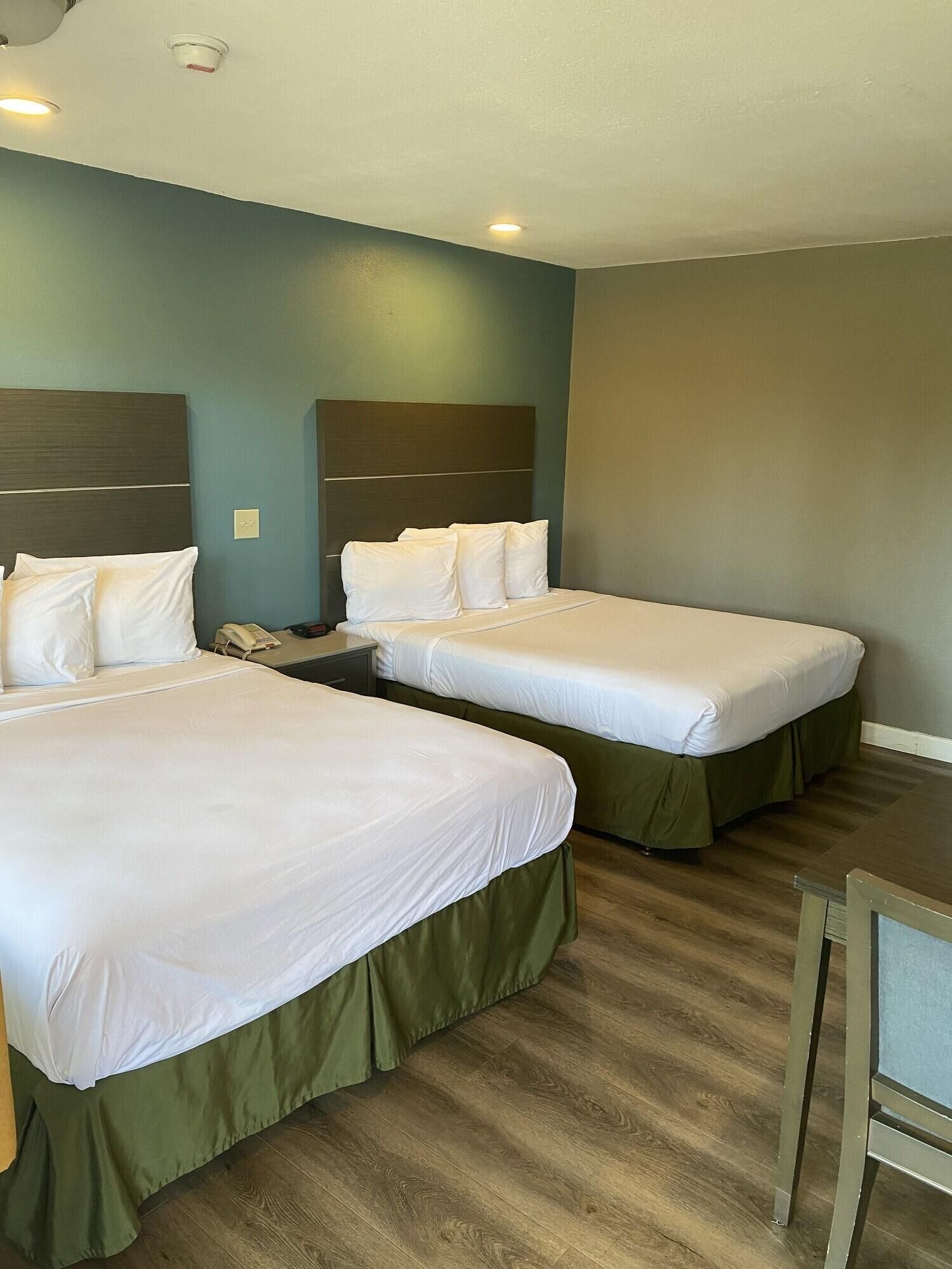 Ein Hotelzimmer mit zwei Betten, jeweils mit weißer Bettwäsche und grünen Bettlaken. Der Raum hat einen Holzboden und hellblaue Wände.