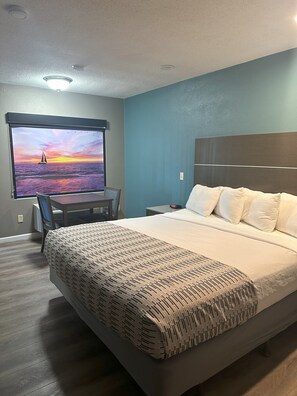 Hotellihuone, jossa on sänky, työpöytä tuolilla, yöpöytä ja seinälle asennettu televisio, josta näkyy auringonlaskumaisema.