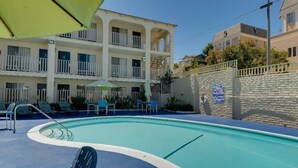 A pool area with lounge chairs and umbrellas, a stone wall with a sign, and a multi-story building with balconies.