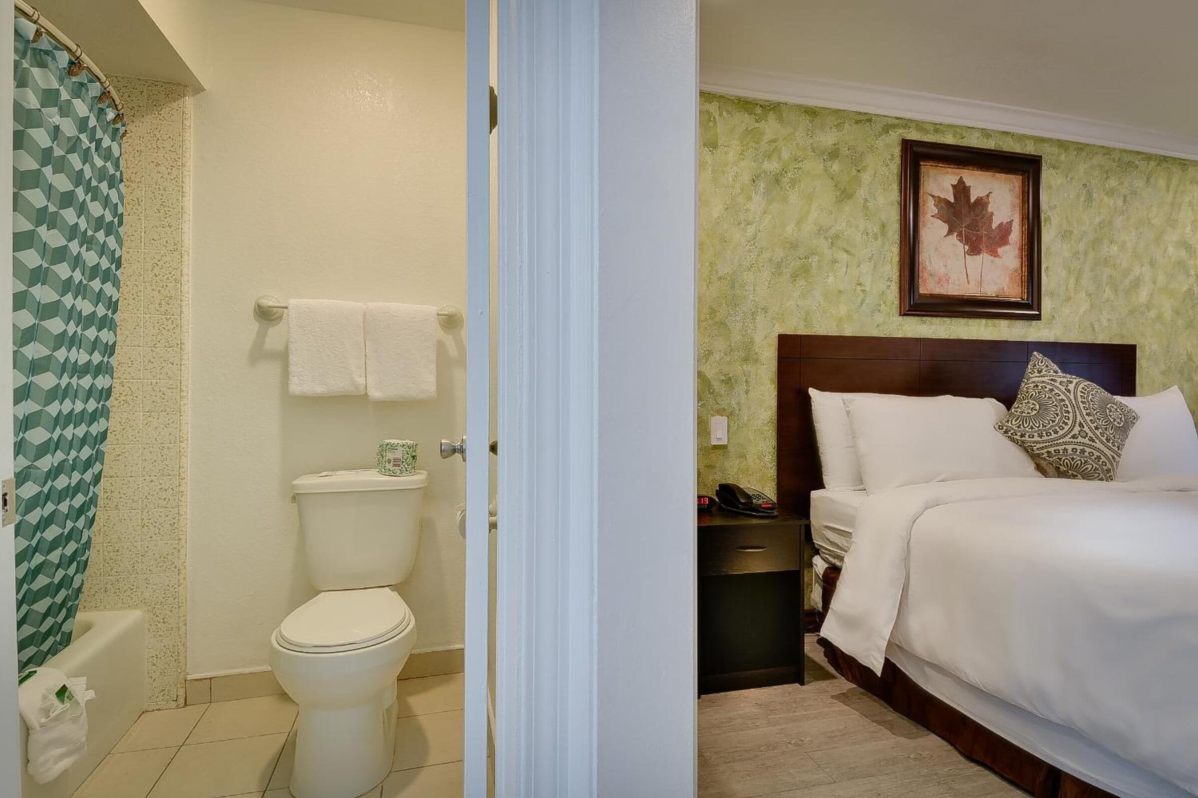 A hotel room with a bathroom featuring a toilet, sink, and shower curtain. The bedroom has a bed with pillows and a framed picture on the wall.