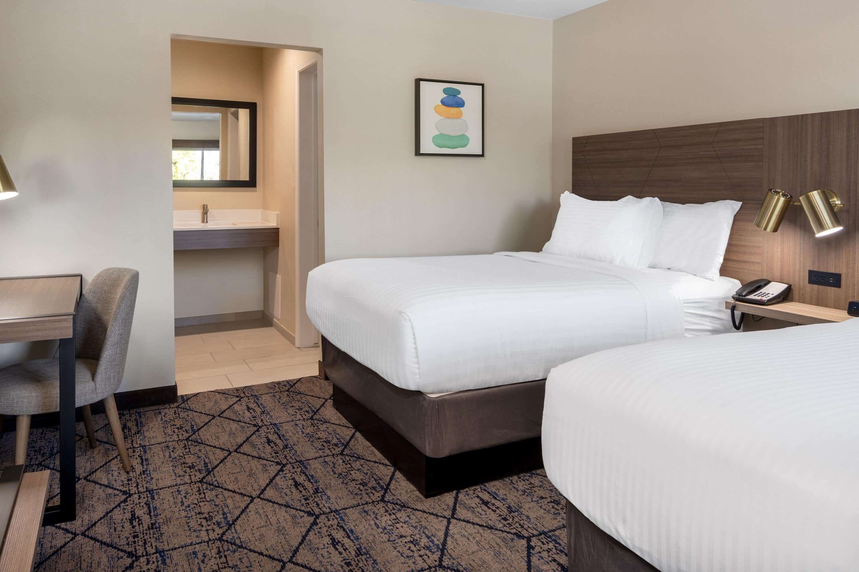 A hotel room with a bed, bedside table, lamp, and a small desk with a chair.